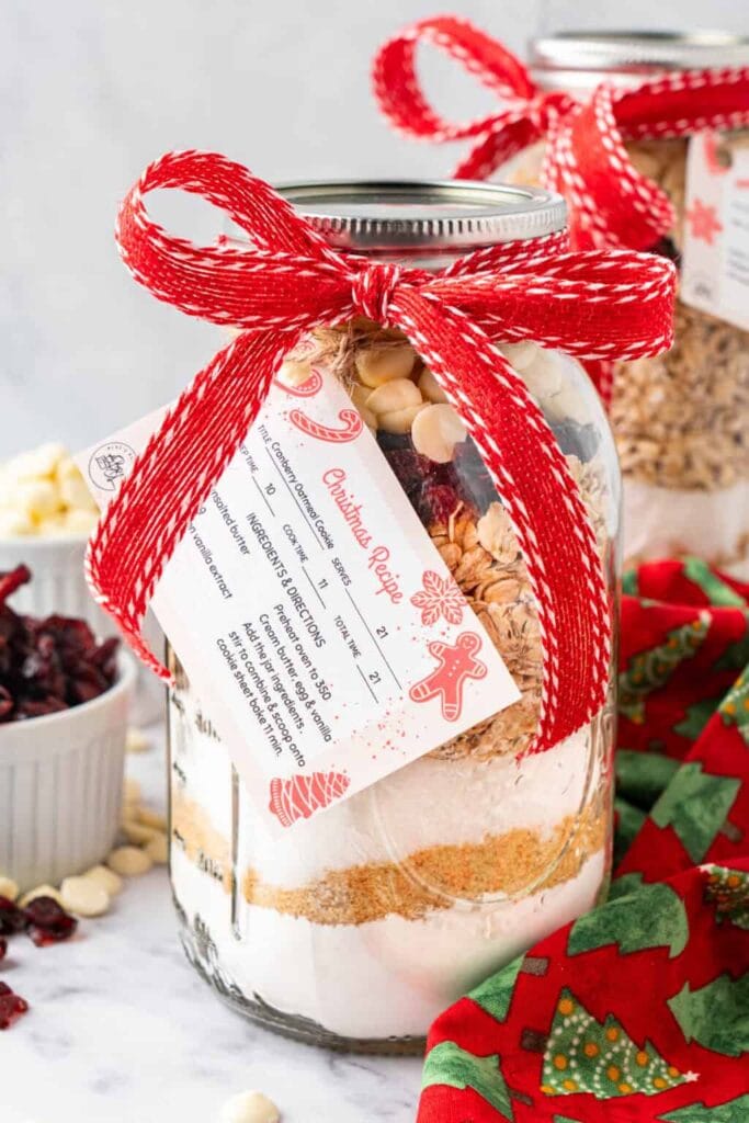 Glass jar layered with flour, sugar, oats, cranberries, and white chocolate chips, tied with ribbon and recipe card, with festive cloth and bowls of ingredients nearby.