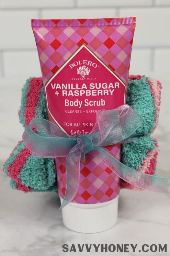 Tube of Bolero Vanilla Sugar + Raspberry body scrub placed in front of rolled pink and teal towels tied with teal ribbon, set against white tile background.