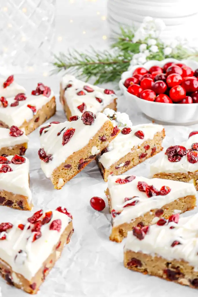 Triangular cranberry dessert bars with white frosting and dried cranberry topping, arranged on white surface beside bowl of cranberries, greenery, and stacked plates.