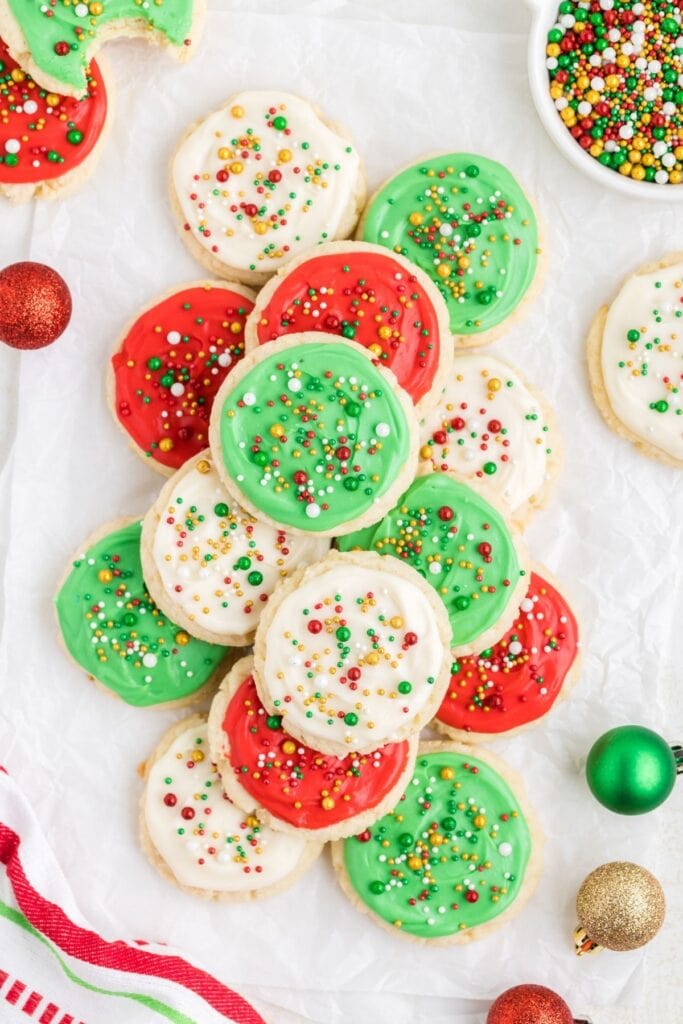 Round sugar cookies frosted in red, green, and white, topped with multicolored sprinkles, arranged on parchment with glittery ornaments and bowl of sprinkles nearby.