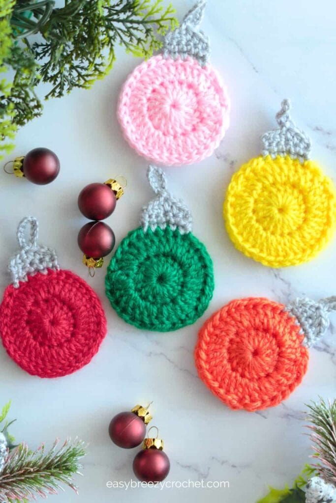 Five colorful crocheted Christmas bauble ornaments with gray tops, arranged with red baubles and pine sprigs on light surface.