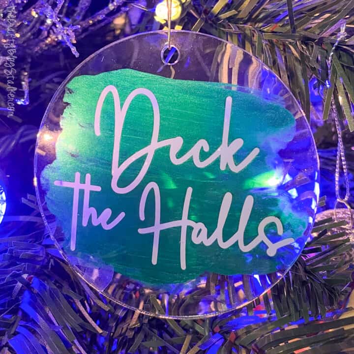 Transparent bauble with green-blue background and white “Deck the Halls” text, hanging on Christmas tree glowing with blue and purple lights.