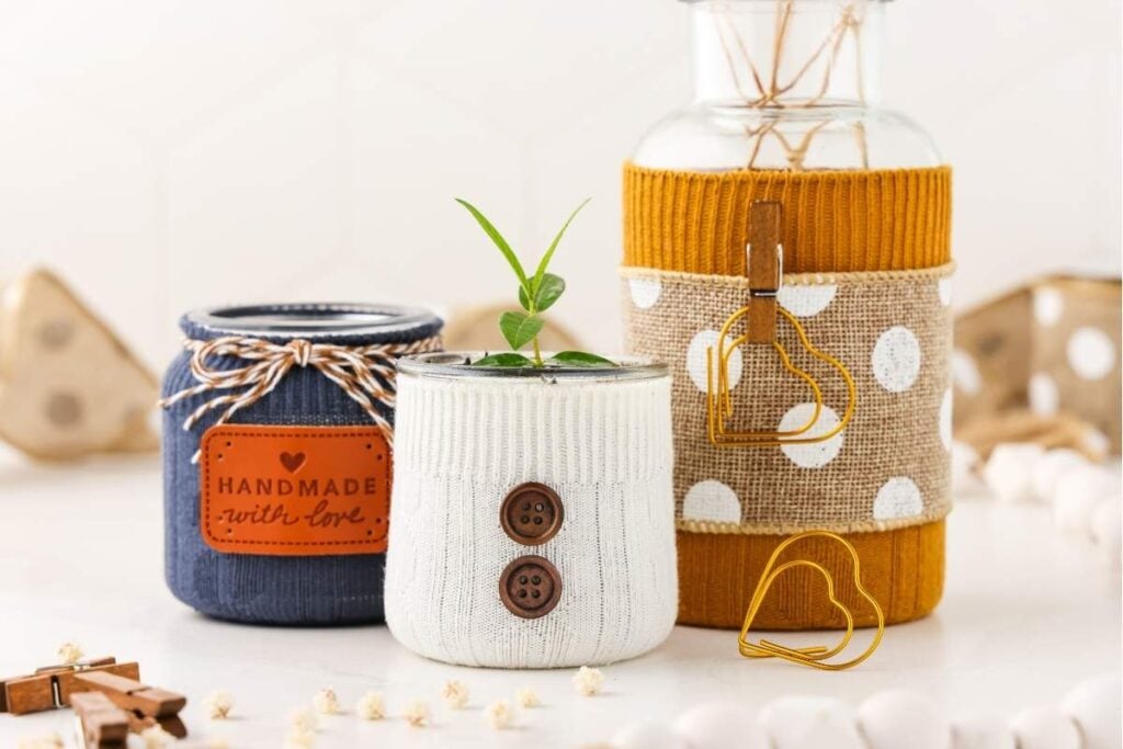 Three fabric-wrapped jars with knit textures, buttons, twine, and heart clip, arranged with clothespins and beads on white surface.