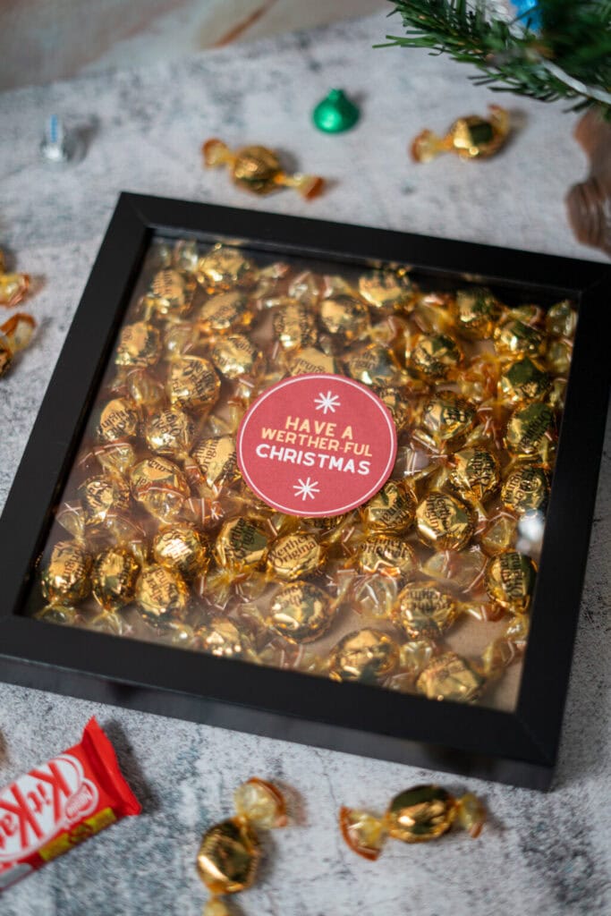 Black-framed box filled with gold-wrapped Werther’s candies, centered red label reading “HAVE A WERTHER-FUL CHRISTMAS,” surrounded by extra candies, KitKat bar, and Christmas tree decor.
