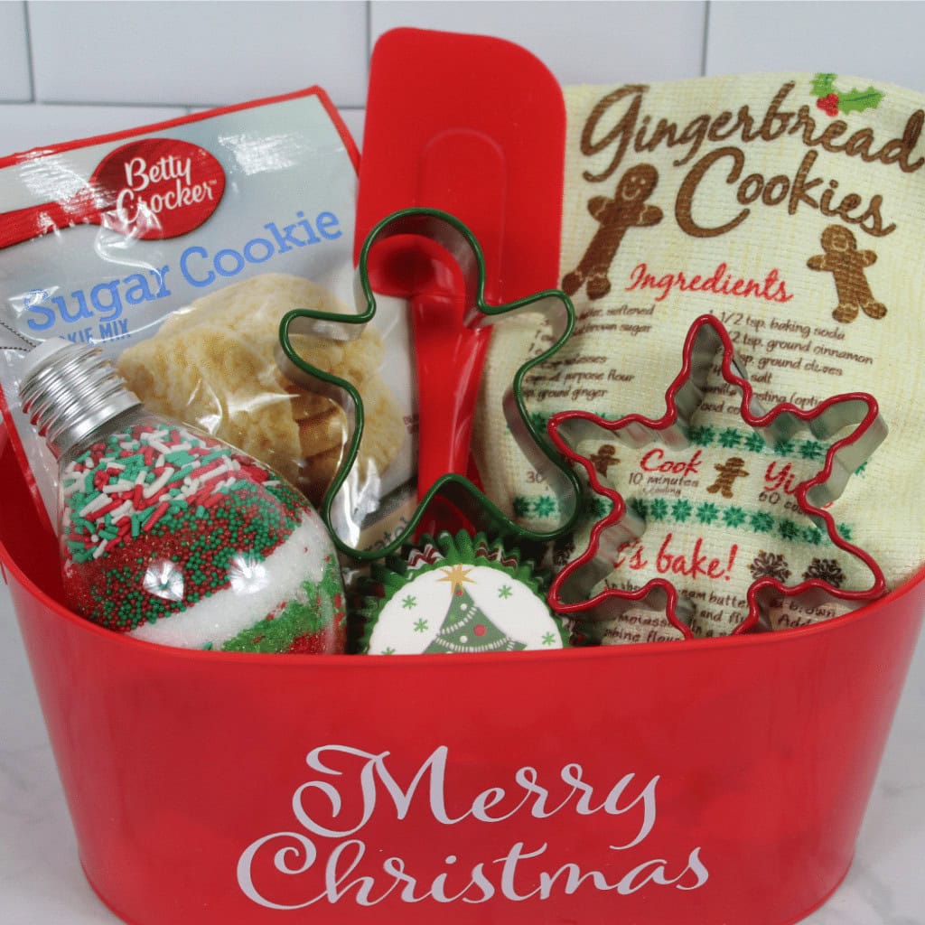 Red container labeled “Merry Christmas” filled with sugar cookie mix, spatula, sprinkles, cookie cutters, cupcake liners, and gingerbread packet.