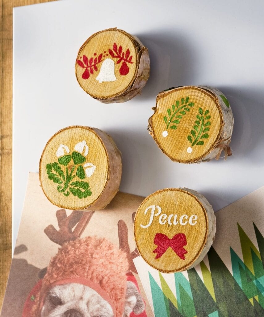 Four wooden slices with painted holiday designs—bell with berries, greenery, flowers, and “Peace” with red bow—arranged on white surface above card showing dog in reindeer antlers.