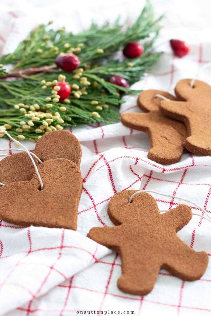 Heart and gingerbread-shaped cinnamon ornaments with hanging loops on white cloth with red grid, surrounded by pine branches and cranberries.