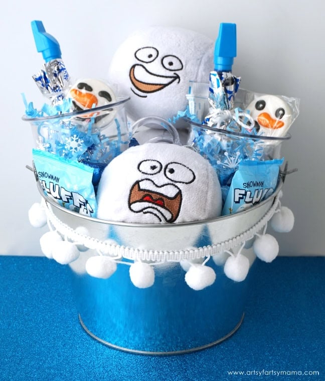 Silver bucket trimmed with pom-poms, filled with plush snowballs, candies labeled “SNOWMAN FLUFF,” snowman treats, and blue party blowers on sparkly blue surface.
