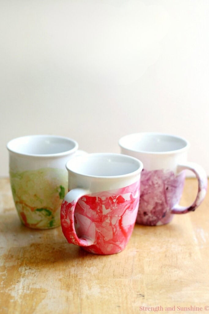Three ceramic mugs with red, purple, and green marbled designs, arranged on wooden surface with plain background.