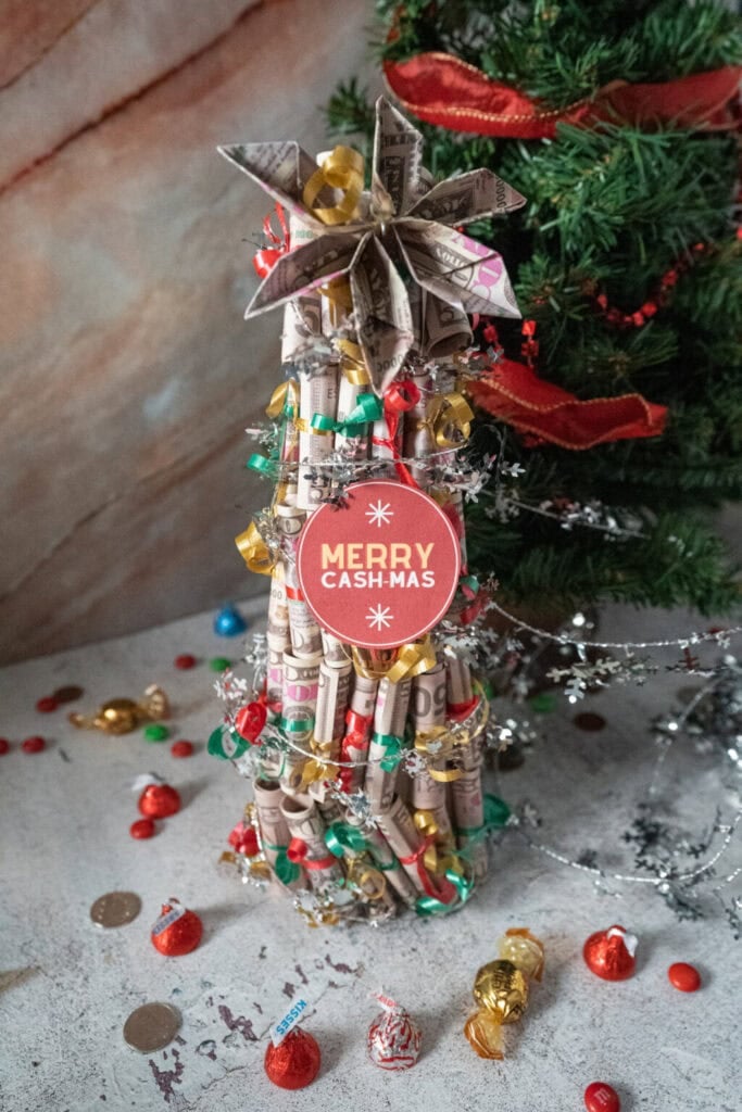 Small Christmas tree made of rolled banknotes with star topper, red tag reading “MERRY CASH-MAS,” surrounded by candies, chocolates, and coins, with decorated tree in background.