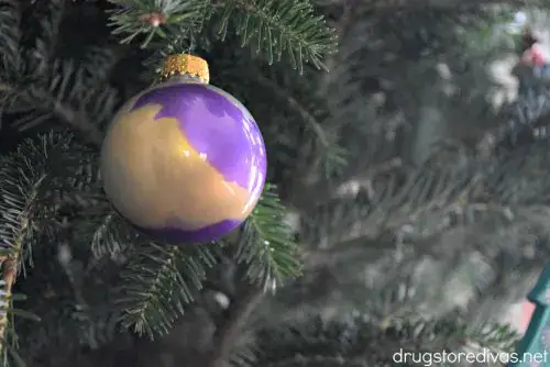 Glossy round Christmas bauble with purple and gold swirl pattern, hanging on pine branch with needles in soft focus.