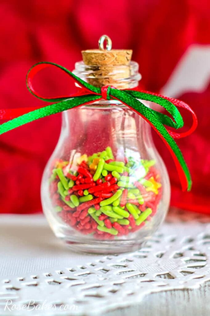 Clear light bulb-shaped jar filled with red and green sprinkles, cork stopper, and ribbon bow, placed on lace surface against bright red background.