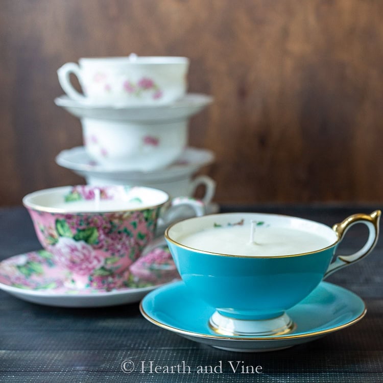 Turquoise and floral teacups repurposed as candles, arranged with stacked vintage cups on dark surface against wooden background.