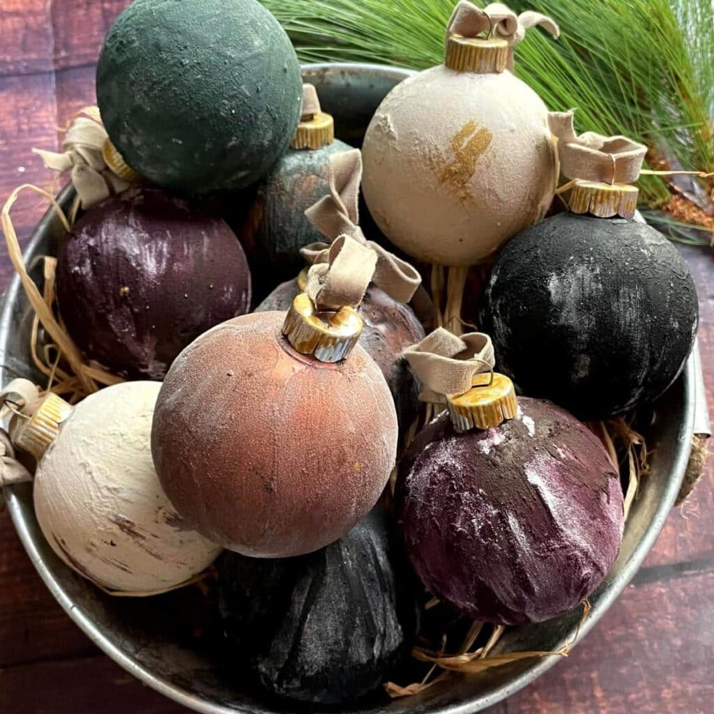 Metal bowl filled with matte textured Christmas baubles in earthy tones, with straw and pine sprigs in background.