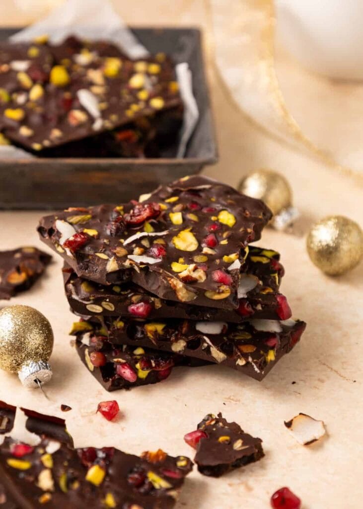 Stack of dark chocolate bark with pistachios, cranberries, pomegranate seeds, and coconut flakes, surrounded by tray of bark and golden ornaments on light surface.