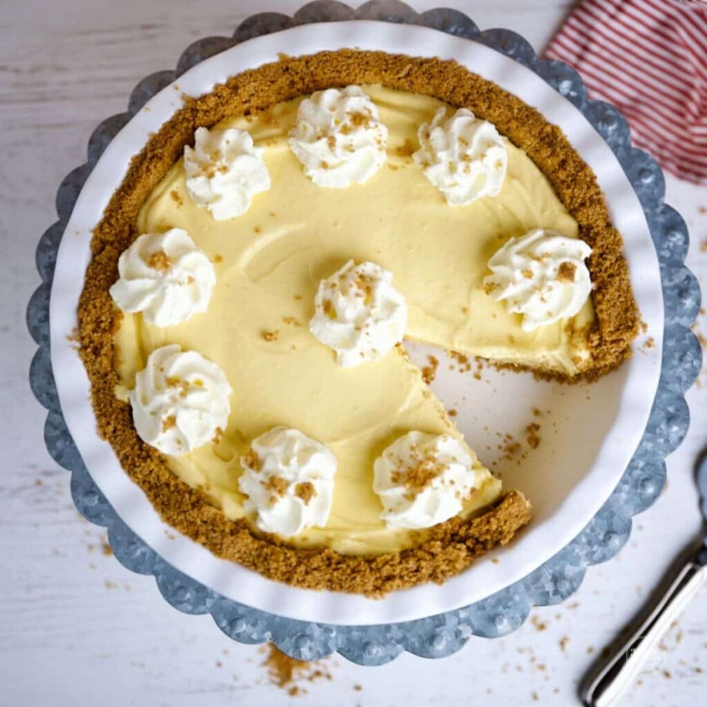 Cream pie with graham crust, pale yellow filling, whipped cream dollops, and sprinkled crumbs, on white plate with striped cloth and pie server nearby.