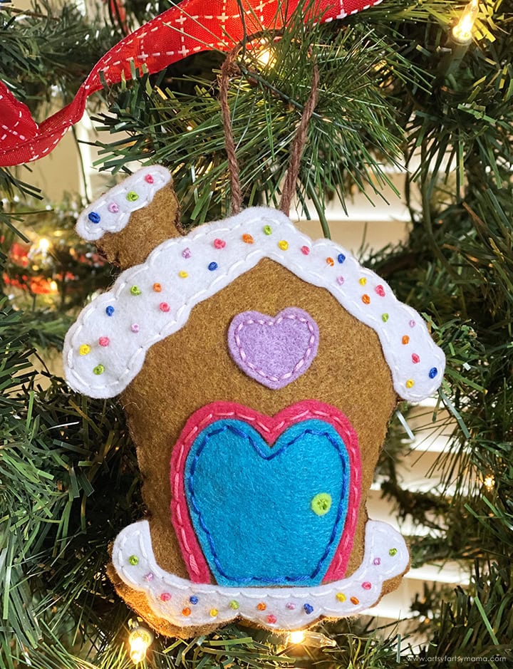Brown felt gingerbread house ornament with white icing trim, colorful bead sprinkles, and blue heart door, hanging on Christmas tree with lights and ribbon.