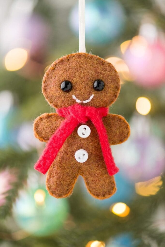 Felt gingerbread man ornament with scarf and stitched smile, hanging on ribbon against blurred Christmas lights and decorations.