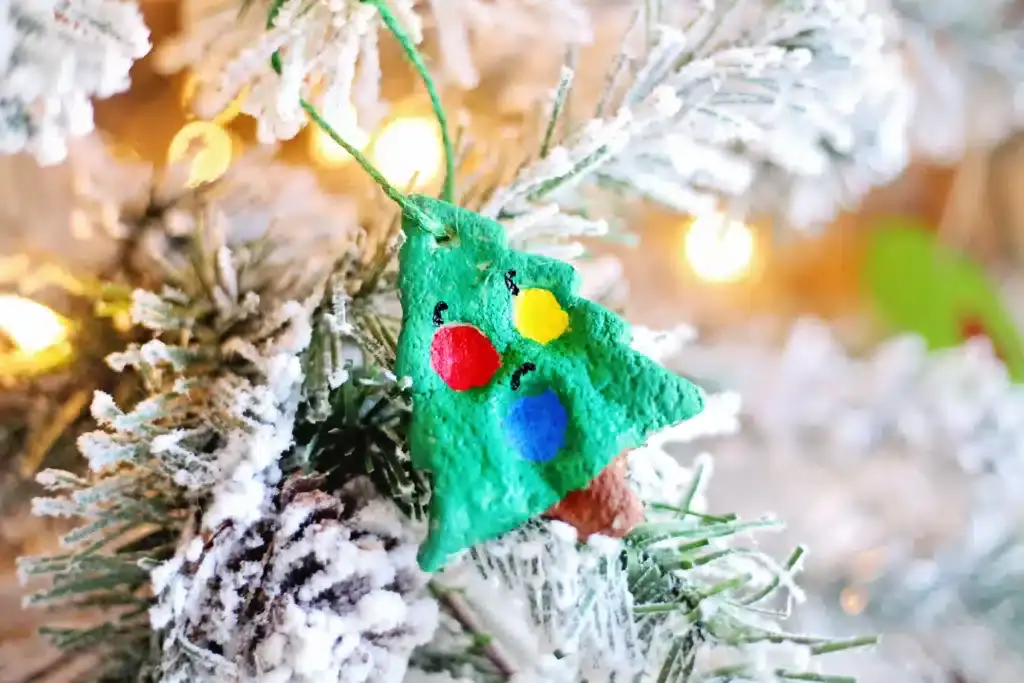Green Christmas tree ornament with colorful circles and face details, hanging on snowy pine branch with lights and pinecone.