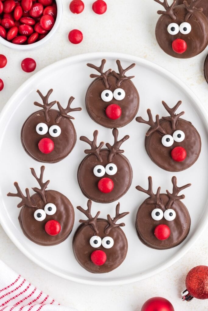 White plate of chocolate-covered cookies decorated as reindeer with candy eyes, red noses, and piped chocolate antlers, surrounded by red candies, ornament, and striped cloth.
