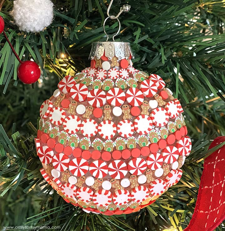 Colorful spherical ornament with peppermint stripes, gingerbread men, and icing-like patterns, hanging on artificial tree with pine needles, berries, and red ribbon.