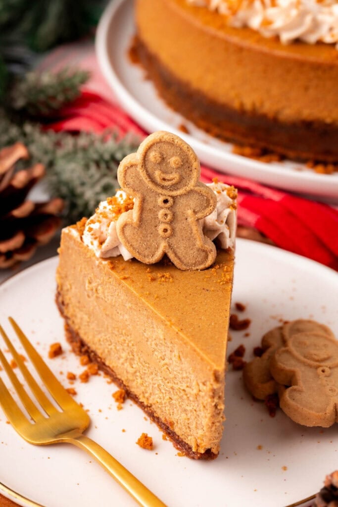 Slice of gingerbread cheesecake with whipped cream and cookie topper, served on white plate with gold fork, surrounded by festive pinecones, greenery, and full cheesecake in background.
