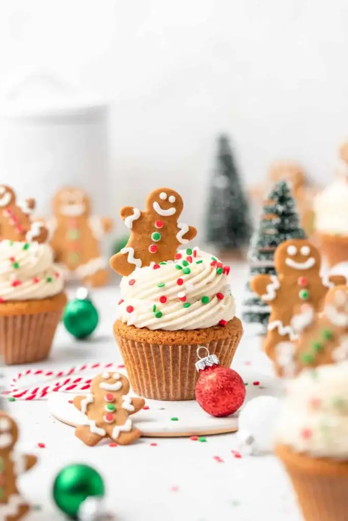 Cupcakes topped with white frosting, sprinkles, and gingerbread cookies, surrounded by ornaments, candy canes, and miniature Christmas trees in festive scene.