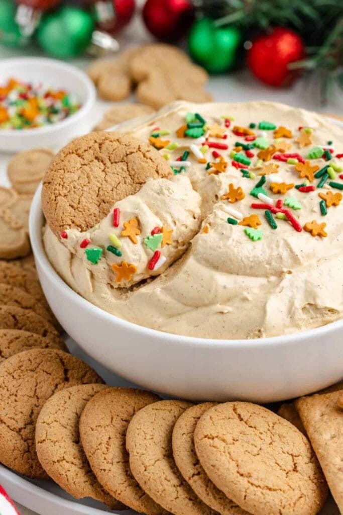 White bowl of creamy dip topped with colorful holiday sprinkles, surrounded by gingersnap cookies, ornaments, and greenery, with one cookie dipped inside.