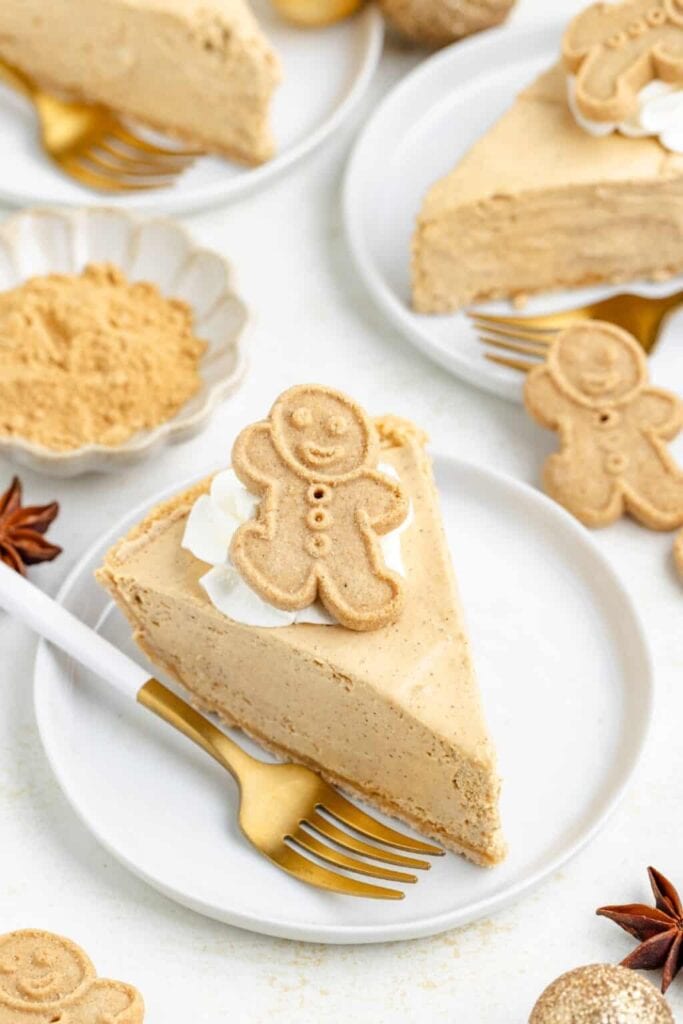 Slice of light brown cheesecake topped with whipped cream and gingerbread man cookie, served on white plate with gold fork, surrounded by cookies, spices, and festive accents.