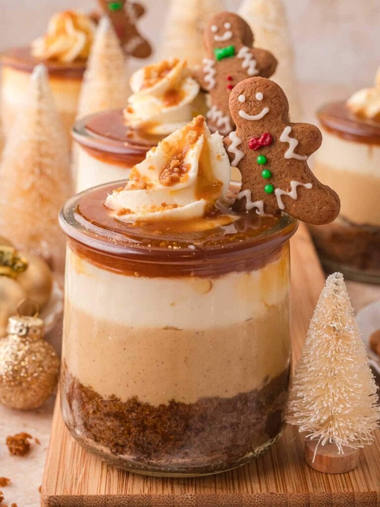 Glass jars layered with spiced cake, cream, and caramel, topped with whipped cream and gingerbread cookies, surrounded by mini Christmas trees and gold ornaments.