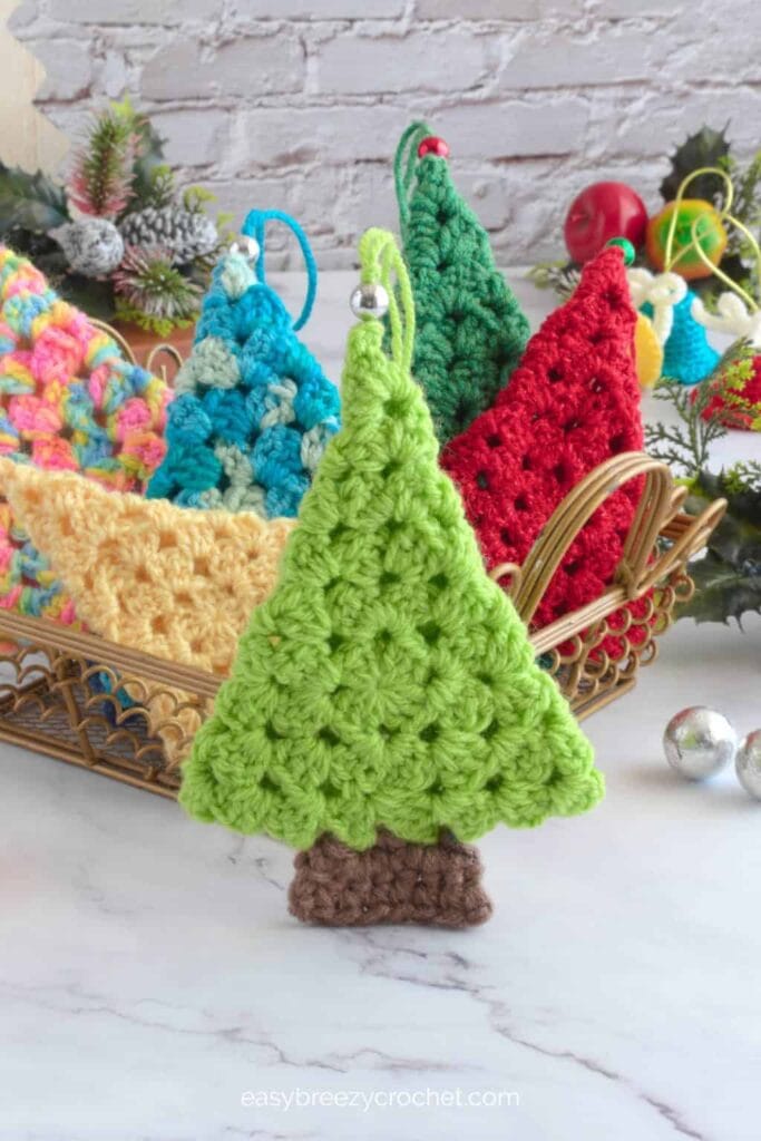Basket of colorful crocheted Christmas tree ornaments with festive decorations and frosted pine branches in background.