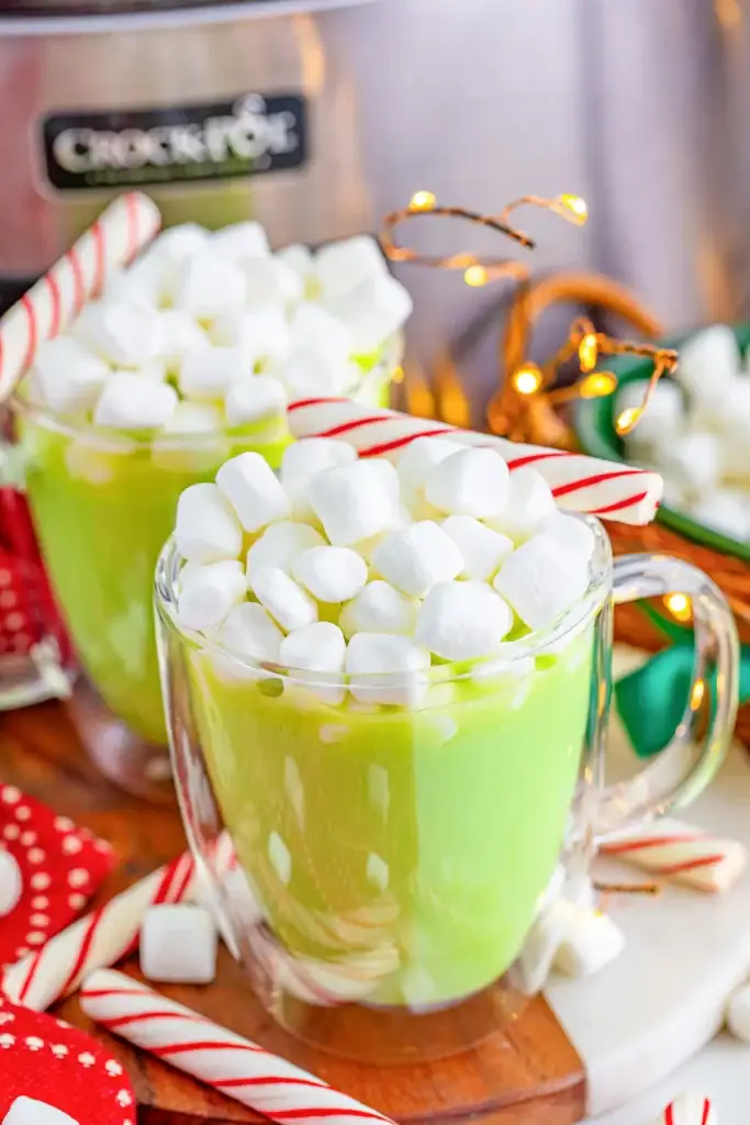 Two glass mugs of bright green drink topped with marshmallows and candy canes, set on wooden surface with scattered sweets, string lights, and Crock-Pot in background.