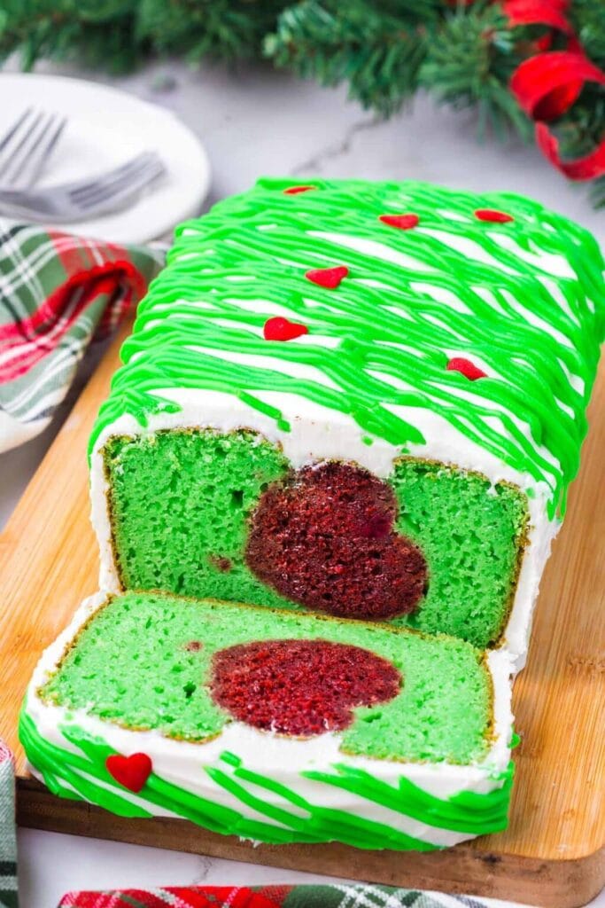 Green loaf cake with red heart center, topped with white frosting, green drizzle, and red heart decorations, displayed on wooden board with plaid cloth and forks nearby.