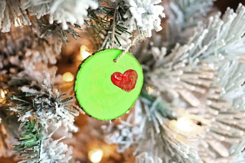 Round green wooden ornament with red heart, hanging on snowy pine branch with warm lights.