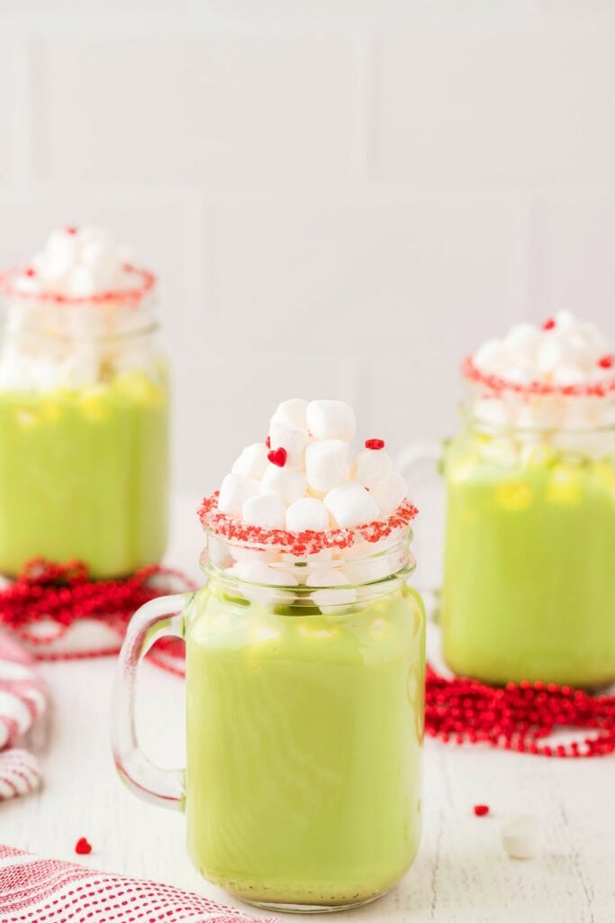Three mason jars filled with light green holiday drink, topped with marshmallows and rimmed with red sugar crystals, arranged on white surface with red beads and striped cloth.