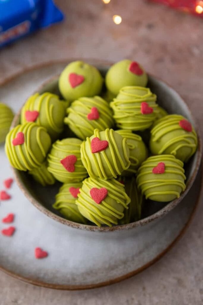 Ceramic bowl of green-coated truffles topped with drizzled icing and red heart candies, set on textured surface with scattered heart decorations.