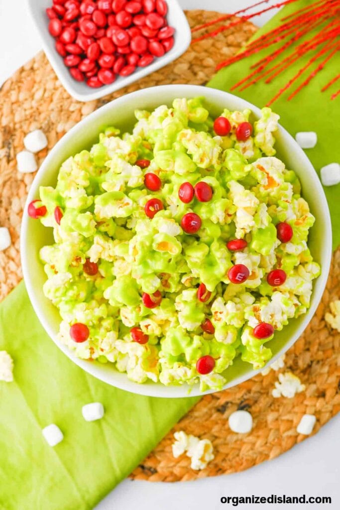 Bowl of bright green candy-coated popcorn topped with red chocolates, set on green cloth with marshmallows, red candies, and holiday decorations nearby.