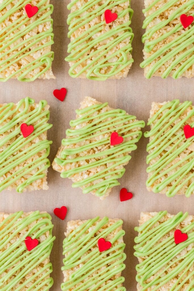 Rectangular Rice Krispies treats drizzled with green icing and topped with small red heart decorations, arranged neatly on parchment surface.