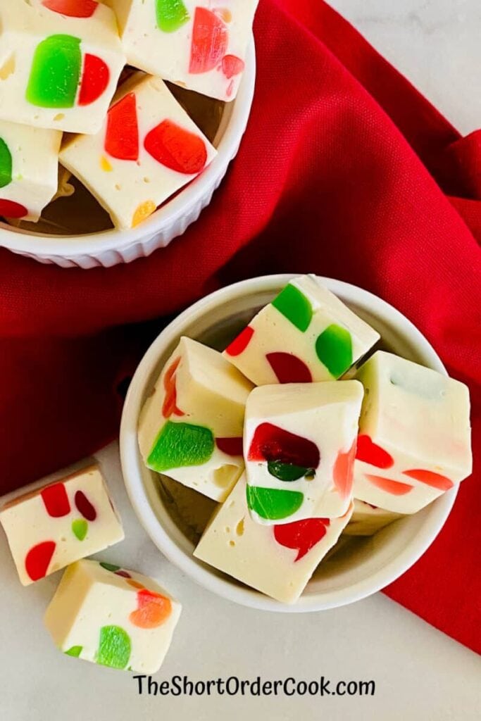 Square pieces of white fudge with red, green, and orange jelly candy chunks, served in white bowls on red cloth background.