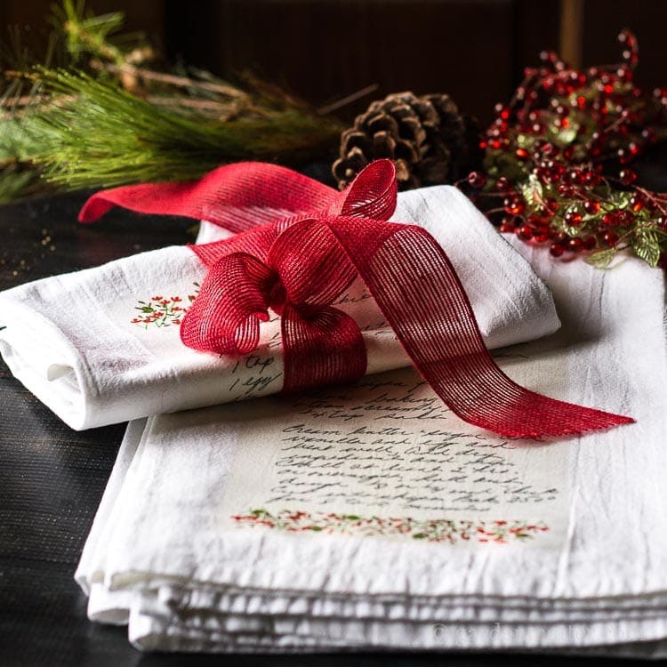 Folded white cloths with handwritten-style print and floral accents, tied with red ribbon, surrounded by pinecones, evergreen, and berries on dark wooden surface.