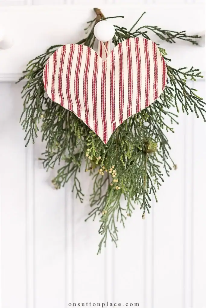 Striped red-and-white fabric heart ornament attached to evergreen sprigs with buds, hanging on white wall hook.