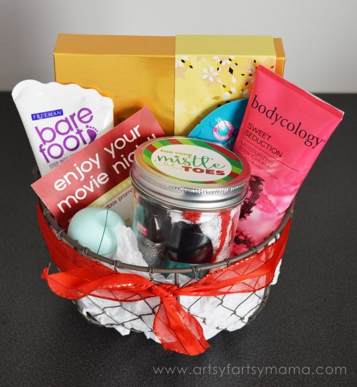 Wire basket lined with tissue and tied with red ribbon, filled with foot cream, body cream, nail polish jar labeled “For your mistle toes,” EOS lip balm, snowflake box, and card reading “enjoy your movie night.”