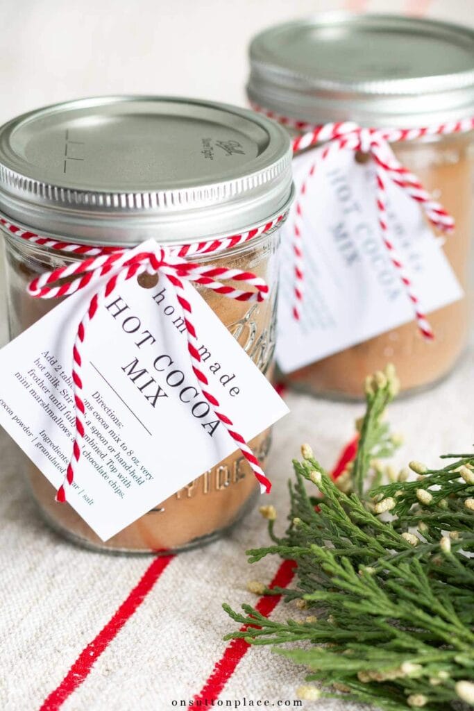 Two glass jars of hot cocoa mix with striped string and recipe tags, placed on striped fabric with pine sprigs.