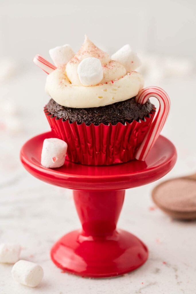 Chocolate cupcake in red wrapper topped with white frosting, red sugar sprinkles, marshmallows, and mini candy cane, displayed on red pedestal with scattered marshmallows nearby.