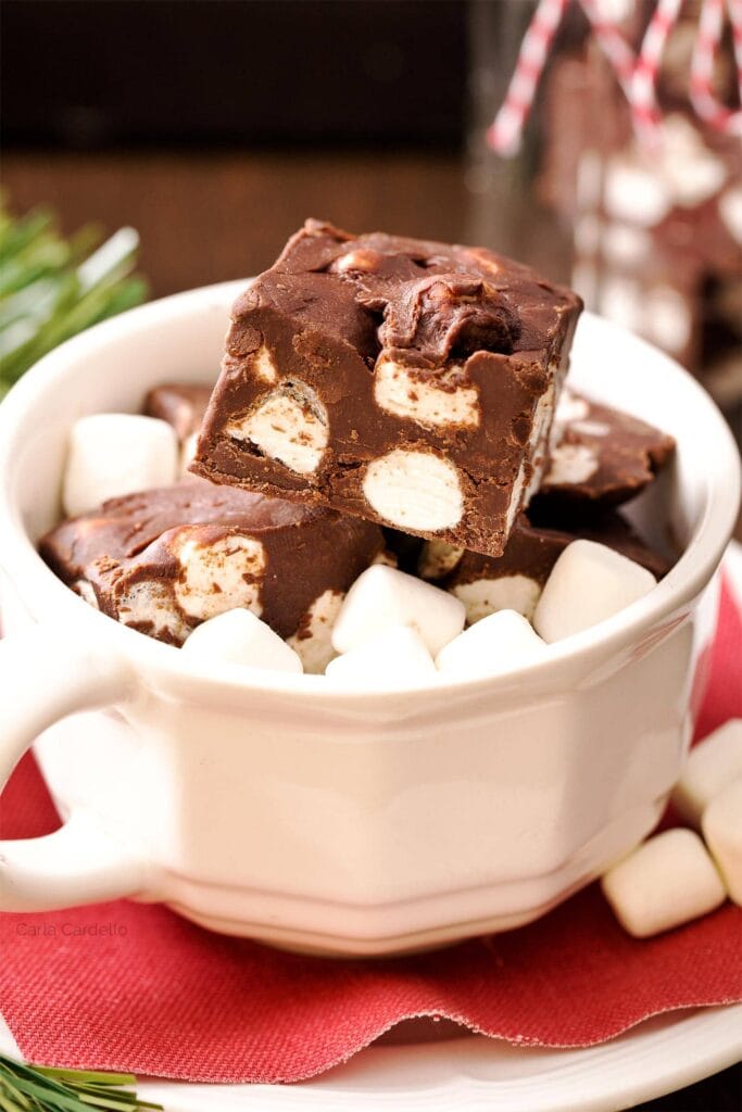 White mug filled with marshmallows and chocolate fudge pieces with embedded marshmallows, set on red napkin and white plate, surrounded by scattered marshmallows and festive décor.