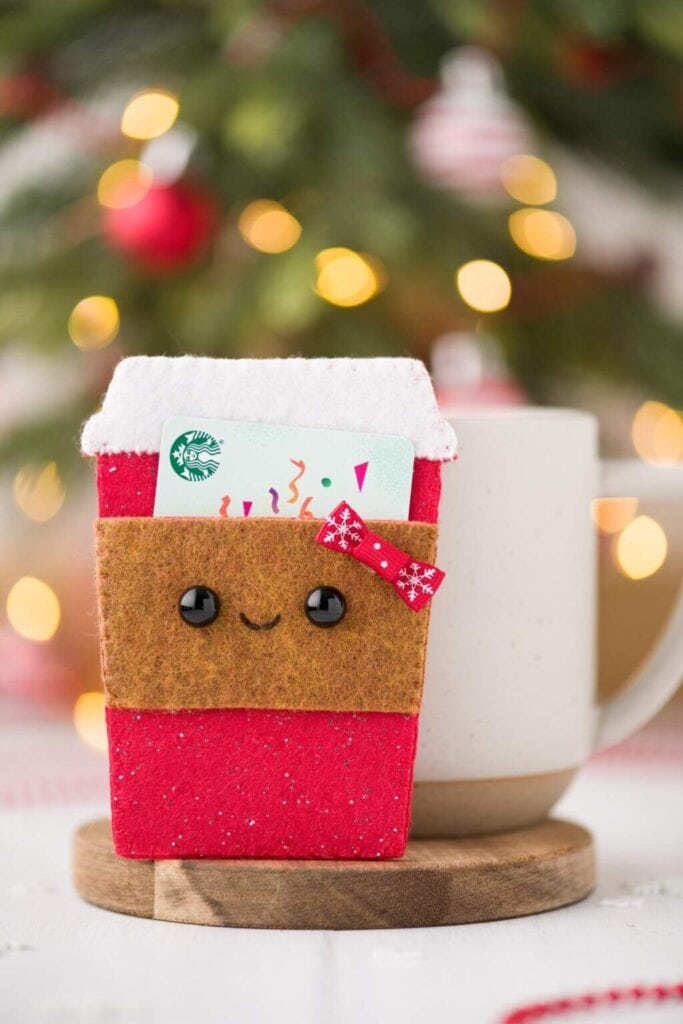 Felt coffee cup gift card holder with red glitter base, smiling brown sleeve, bow, and visible Starbucks card, set against blurred Christmas tree background.