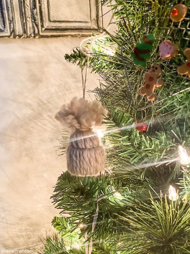 Beige yarn winter hat ornament with pom-pom, hanging on Christmas tree with lights, gingerbread figures, and festive decorations.
