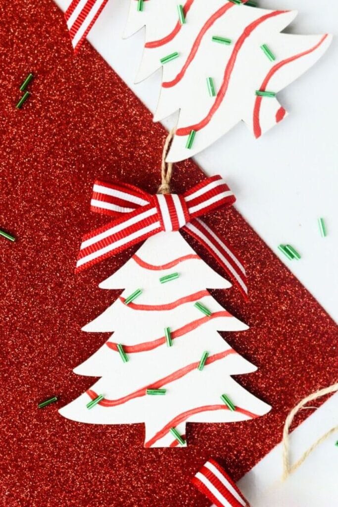White Christmas tree ornament with red wavy lines and green sprinkle-like beads, tied with striped ribbon bow and twine loop, set against red glittery and white background.