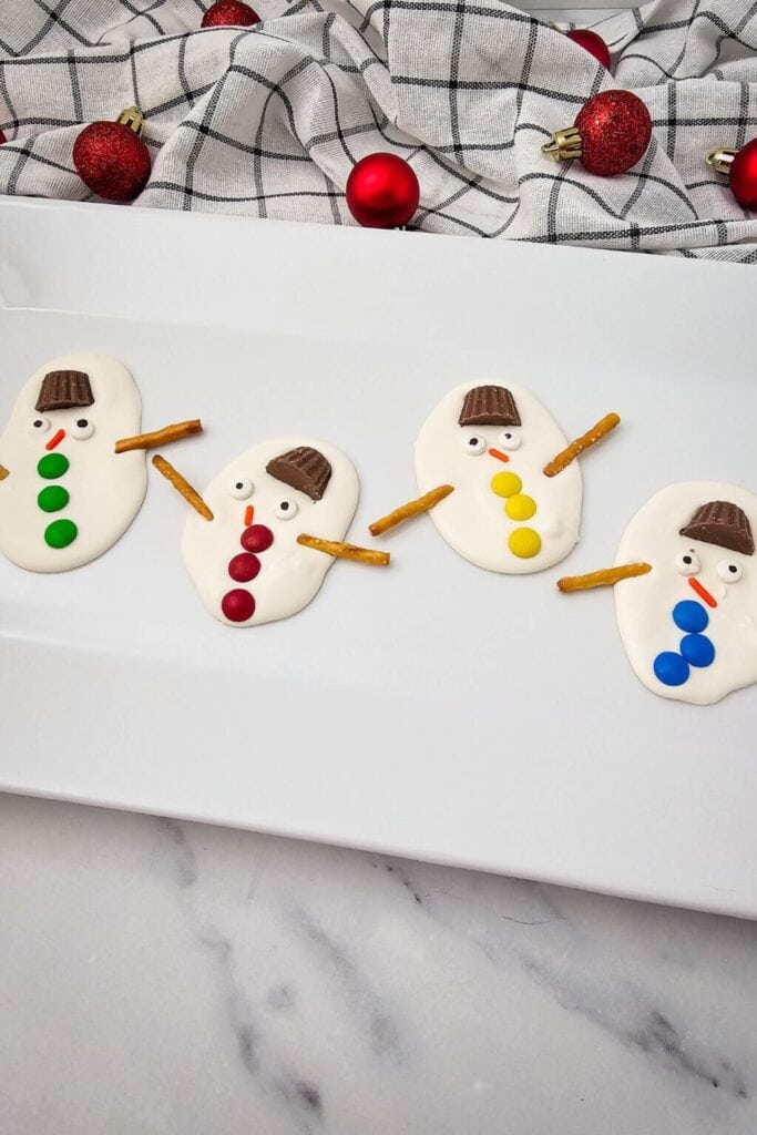 Snowman-shaped treats with candy hats, eyes, noses, and buttons, plus pretzel arms, arranged on white plate with festive ornaments in background.