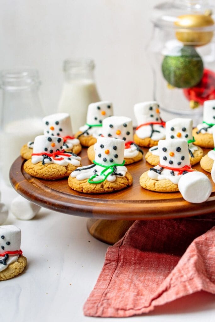 Cookies decorated as melting snowmen with marshmallow heads, icing snow, and colorful scarves, arranged on wooden cake stand with milk bottles, ornaments, and red napkin.